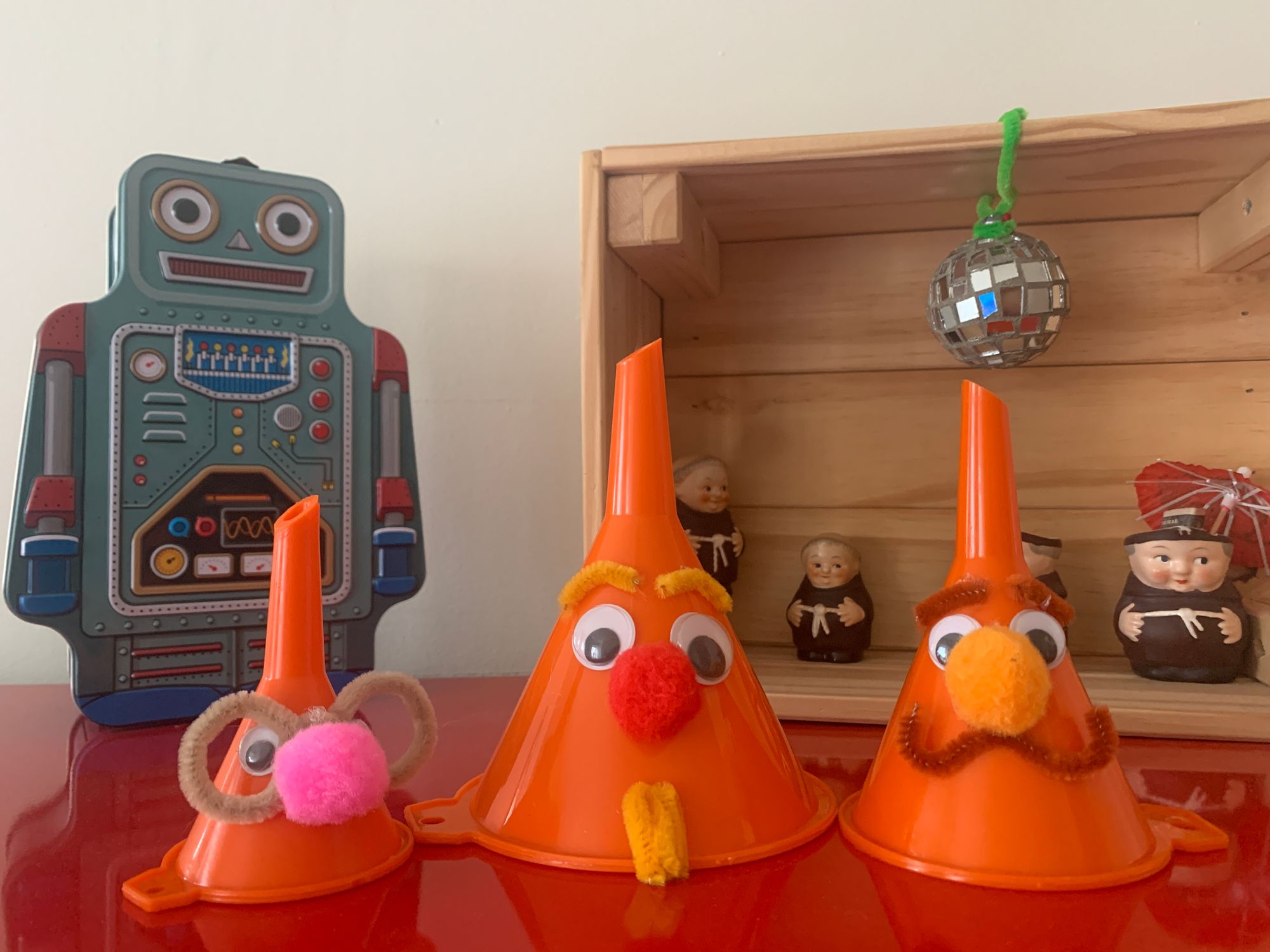 Three funnels upside down decorated with googly eyes, pom-poms, and pipe cleaners to look like people. Behind them is a lunch box robot and a small diorama in a crate with a bunch of small ceramic friars and a disco ball.